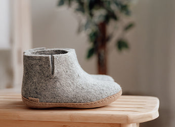 A junior glerups boot in the color Grey is placed on a wooden table indoors, with soft natural light and blurred greenery in the background.