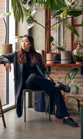 A person sits indoors in a modern room with plants and large windows, wearing dark clothing and Charcoal grey colored glerups Langdal sneakers.