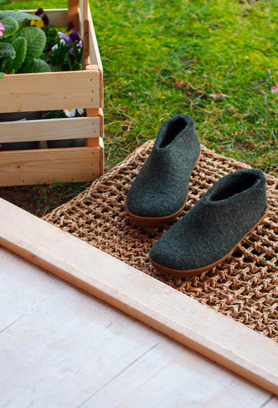 A pair of glerups shoes in the color Forest green placed on a woven doormat next to a wooden crate with potted plants. The shoes are positioned outdoors on a surface where the doormat meets grass.