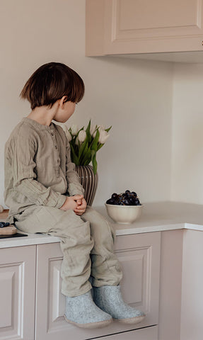 A child sitting on a kitchen counter wearing glerups junior boots in the color Grey, with a bowl of dark berries and a vase of green plants beside them.